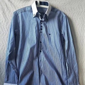 Blåvit randig skjorta från Armani.W50/L70 - Snygg blåvit randig skjorta från Armani med klassisk vit krage och långa ärmar. Skjortan har svarta knappar och ett diskret broderat logomärke på bröstet. Perfekt för dig som gillar stilrena detaljer och tidlös design.
