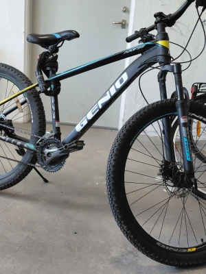 Svart och blå mountainbike Genio 27,5  - Säljer en svart mountainbike från Genio med blå och gula detaljer. Cykeln har grova däck, skivbromsar och sportig ram. Perfekt för dig som gillar att cykla i terräng eller på grusvägar. Sadel och handtag i svart, samt växlar och stötdämpare fram.