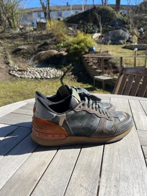 Valentino Camo Sneakers med nitar - Snygga sneakers från Valentino med kamouflagemönster i grönt och svart, detaljer i grått och orange mocka samt coola nitar på hälen. Skorna har svarta skosnören och en klassisk låg siluett. Perfekta för dig som gillar streetstyle och exklusiva märken.