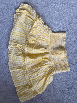 Gul rutig volangkjol - Söt kort kjol i gult och vitt med rutigt mönster och volang. Kjolen har en bred, smockad midja som ger en bekväm och snygg passform. Perfekt för varma dagar och somriga outfits.