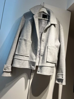 Ljusgrå jacka med stora fickor - Snygg ljusgrå jacka från COS i ullmix med rak, boxig passform. Jackan har stora utanpåliggande fickor, bred krage och knappar framtill samt vid ärmslut. Perfekt för lager på lager och en clean look. Storlek XL men jag har S/M i vanliga fall och har haft denna som oversize. 
