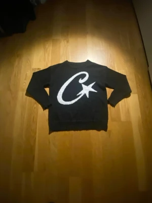 Corteiz stickad tröja  - Cool svart stickad tröja med ett stort vitt C och en stjärna på bröstet. Tröjan har rund halsringning och långa ärmar. Perfekt för dig som gillar grafiska mönster och vill sticka ut lite extra.