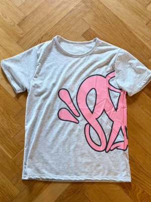 Grå t-shirt med rosa tryck - Snygg grå t-shirt med ett stort rosa grafiskt tryck på framsidan. T-shirten har rund halsringning och korta ärmar. Perfekt för en avslappnad och trendig stil.