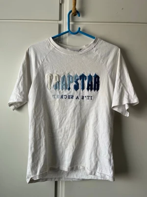 Vit Trapstar t-shirt med blått tryck medföljer shorts - Säljer en vit t-shirt och shorts set från Trapstar med ikoniskt broderat Trapstar-tryck i blå och vita nyanser på bröstet samt texten 'It's a secret' under. Klassisk rund hals och korta ärmar. Tillverkad i mjuk bomull för skön passform.