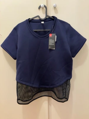 Mörkblå croppad topp från Under Armour - Sportig mörkblå croppad topp från Under Armour med korta ärmar och meshdetalj nedtill. Perfekt för träning eller en aktiv vardag. T-shirten har rund halsringning och en modern, loose passform.