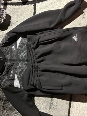 Svart träningsset från Adidas - Säljer ett svart träningsset från Adidas med jacka och byxor. Jackan har dragkedja, hög krage och mönstrade detaljer i vitt och grått upptill. Byxorna har resår i midjan och Adidas-logga på benet. Perfekt för träning eller chill.