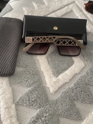 Svarta solglasögon från Givenchy - Stilrena solglasögon från Givenchy med fyrkantiga svarta bågar och gråtonade linser. Sidorna har ett unikt geometriskt mönster i beige och metall med Givenchy-logga. Perfekt accessoar för att höja din look.