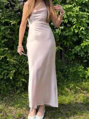 Ljusrosa satinklänning med blommor - Andcänt till lärarmiddagen en gång ❤️ perfekt till bal eller lärarmidag och i gott skick! Säljer en lång, ljusrosa satinklänning med smala axelband och draperad urringning. Klänningen har dekorativa blommor längs banden på ryggen och en elegant, figurnära passform. Perfekt för dig som vill ha en romantisk och stilren look. Från Gina🫰