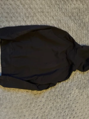 Svart hoodie från GANT med logga - Svart hoodie från GANT i mjuk bomullsblandning. Klassisk modell med huva och dragsko, samt broderad GANT-logga och sköld på bröstet. Insidan av huvan har grått tyg och en randig detalj. Perfekt för en avslappnad och stilren look.