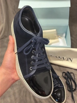 Lanvin sneakers i blå mocka och lack - Snygga sneakers från Lanvin i mörkblå mocka med svart lackad tå och vita sulor. Klassisk låg modell med blå skosnören och extra par snören medföljer. Kommer med original ask och dustbag. Perfekta för dig som vill ha en stilren och lyxig look.