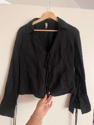 Svart blus med knytning framtill - Trendig svart blus med långa ärmar och knytning framtill. Den är linne material från Zara. Använd fåtal gånger och hängt i garderoben, därav lite skrynklig, så kan behöva strykning.