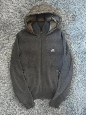 Moncler cardigan - Snygg grå stickad jacka från Moncler med quiltad huva i blankt material.                       Extremt eftertraktad moncler cardigan.  I riktigt bra skick. Självklart äkta, har kvitto. Endast seriösa köpare.  Skriv vid fler frågor, storleken är L men passar M