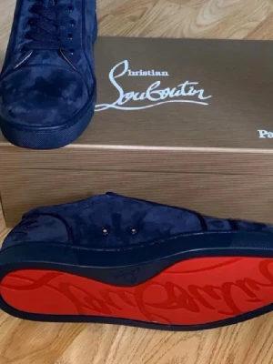 Marinblå mockasneakers från Louboutin - Snygga marinblå sneakers från Christian Louboutin i mjuk mocka med ikonisk röd sula och broderad logga på hälen. Skorna har en stilren design med matchande marinblå snörning och detaljer. Perfekta för dig som vill sticka ut med exklusiv känsla.