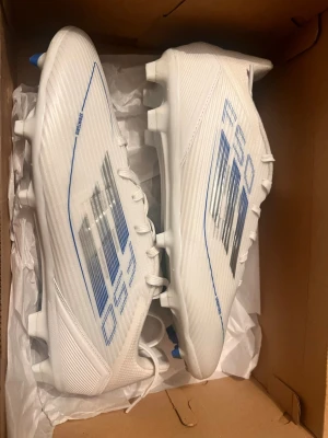 Adidas F50 fotbollsskor - Säljer ett par vita Adidas F50 fotbollsskor med blå och svarta detaljer. Skorna har snörning, låg profil och är tillverkade i ett lätt syntetmaterial för bästa känsla och fart på planen. Perfekta för dig som vill ha snabba och stilrena skor.