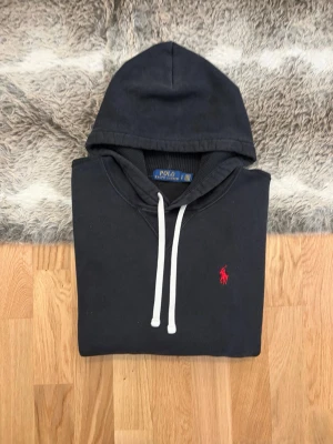 Svart Polo Ralph Lauren Hoodie - Storlek S - Hej! 👋 Säljer en stilren svart Polo Ralph Lauren hoodie i storlek S (Herr). Den har den klassiska röda pony-loggan broderad på bröstet 🐎 Tröjan är i bra skick och har den sköna mjuka fleece-insidan kvar ✨ Det kan förekomma en naturlig, lätt solblekning på vissa ställen, vilket ger den en snygg vintage-känsla 👕 Sitter perfekt (Modell: 177cm). Modellen saknar QR-kod men alla tvättlappar sitter kvar ✅ Ett oumbärligt basplagg som passar till allt! Fraktar snabbt 🚛📦💬💯 