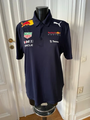 Red Bull Racing pikétröja Puma - Mörkblå pikétröja från Puma med Red Bull Racing-loggor och flera sponsorsloggor på bröstet och ryggen. Tröjan har krage med knappar och korta ärmar. Perfekt för motorsportfans som vill sticka ut med en sportig och snygg look.