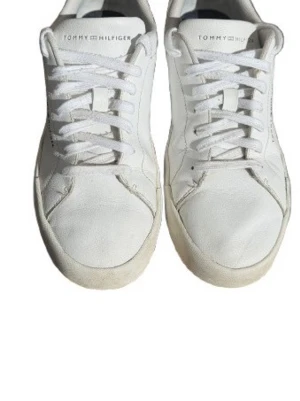 Vita sneakers från Tommy Hilfiger - Klassiska vita sneakers från Tommy Hilfiger med lågt skaft och vita snören. Skorna har diskret logga på sidan och mörkblå detalj vid hälen. Tillverkade i slätt läder med stilren design som passar till det mesta.