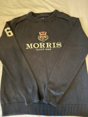 Mörkblå stickad tröja från Morris - Mörkblå stickad tröja från Morris med broderad logga och brittisk flagga på bröstet samt siffran 6 på ena ärmen. Tröjan har rund halsringning och ribbade muddar vid ärmslut och nederkant. Perfekt för en klassisk och stilren look.