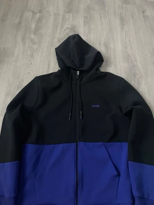 BOSS saggy1 - Snygg hoodie från BOSS i svart och blått med dragkedja framtill och två fickor. Huva med dragsko och diskret BOSS-logga på bröstet. Tillverkad i mjuk bomullsblandning för skön känsla. Perfekt för en avslappnad streetwear-look.