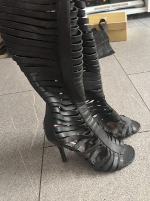 Svarta höga pumps med remdetaljer - Unika svarta pumps med höga klackar och coola remdetaljer som går upp till knäna. Skorna har öppen tå och dragkedja baktill för enkel påtagning. Tillverkade i konstläder med edgy design som sticker ut.