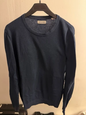 Mörkblå tröja från Jack & Jones - En stilren mörkblå tröja från Jack & Jones med rund halsringning och långa ärmar. Tröjan är tunnstickad och har en klassisk passform som funkar till många olika outfits. Perfekt för dig som gillar enkel och snygg stil.