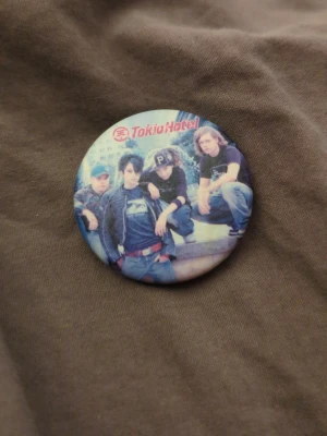 Tokio Hotel pin med bandfoto - Cool pin med motiv av bandet Tokio Hotel. Rund form med färgglatt tryck där bandmedlemmarna syns och loggan står högst upp. Perfekt accessoar för dig som gillar bandmerch och vill visa din stil.