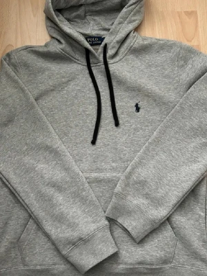 Grå Polo Ralph Lauren - Snygg grå hoodie från Polo Ralph Lauren med svart dragsko och klassisk broderad logga på bröstet. Mjuk bomullsblandning och känguruficka. Perfekt för en chill och stilren look. Passar perfekt året om. Storlek S men passar även M