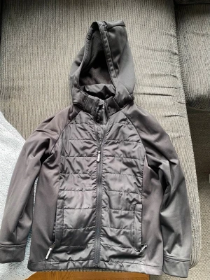 Mörkgrå vindjacka med huva - Snygg mörkgrå vindjacka med quiltade partier framtill och baktill. Jackan har dragkedja, två fickor med dragkedja och en skön huva. Materialet är en mix av slät och vadderad textil, perfekt för blåsiga dagar.