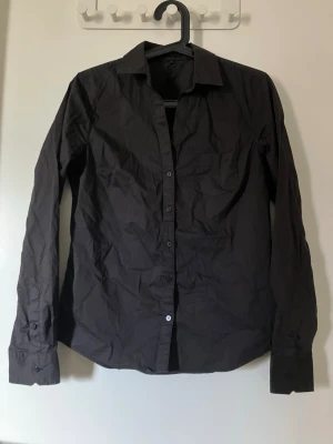 Svart klassisk skjorta med knappar - En stilren svart skjorta med lång ärm och knappar framtill. Skjortan har klassisk krage och bröstficka, perfekt för en clean och enkel look. Tillverkad i bomull för en bekväm känsla.