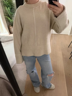 Beige stickad tröja från Zara - Mysig beige stickad tröja från Zara med rund halsringning och breda ribbade muddar vid ärmslut och nederkant. Tröjan har en dekorativ söm framtill och en relaxed, loose passform som passar perfekt till jeans.