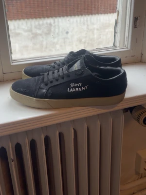 Svarta Saint Laurent sneakers - Svarta sneakers från Saint Laurent med vit logga på sidan och vit sula. Klassisk låg modell med svarta skosnören och detaljer i läder och textil. Perfekta för en stilren och trendig look.