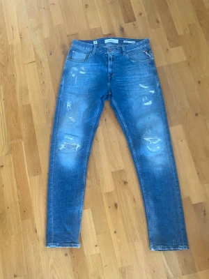 Blå slitna jeans från Replay - Säljer ett par blå jeans från Replay med slitna detaljer på benen. Modell Mickym och slim tapered fit. Storlek W 30 och L 30.