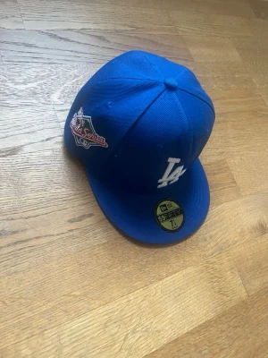 Blå LA Dodgers keps från New Era - Snygg blå keps från New Era med LA Dodgers-logga framtill och World Series-märke på sidan. Modellen är 59FIFTY med platt skärm och klassisk passform. Perfekt för dig som gillar sport och streetstyle.