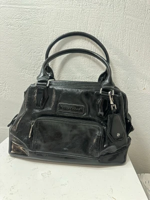 Svart lackad handväska Longchamp - eautiful Longchamp Légende handbag. Glossy black leather, exterior in excellent condition, with a small stain on the bottom of the interior. Metal “feet” on the base. Inside, a spacious zippered side pocket, two smaller pockets, and a leather strap with a clip for keys/keyring. In addition, there is a small exterior zippered side pocket. Dimensions: 30 × 13 cm.