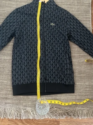 Svart mönstrad ziptröja från Lacoste - Snygg svart ziptröja från Lacoste med diskret rutmönster och klassisk krokodil-logga på bröstet. Tröjan har hel dragkedja, ståkrage och muddar vid ärmslut och nederkant. Perfekt för en sportig och stilren look. Priset kan diskuteras vid snabb äffär 