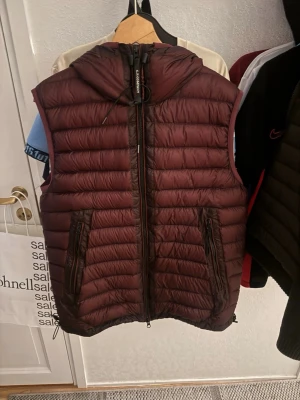 Vinröd dunväst med huva från C.P. Company - Snygg vinröd dunväst från C.P. Company med huva och ikoniska glasögon-detaljer. Västen har två dragkedjeförsedda fickor framtill och justerbar dragsko nertill. Tillverkad i lätt, quiltat material som håller värmen utan att bli klumpig.