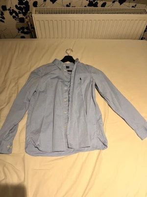 Ljusblå skjorta från Polo Ralph Lauren - Klassisk ljusblå skjorta från Polo Ralph Lauren med button-down krage och broderad logga på bröstet. Skjortan har långa ärmar, knappar framtill och är tillverkad i bomull. Perfekt för en stilren och avslappnad look.