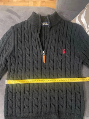 Svart kabelstickad tröja Polo Ralph Lauren - Kabelstickad svart tröja från Polo Ralph Lauren med halv dragkedja och hög krage. Tröjan har ett rött broderat logotyp på bröstet och ribbade muddar vid ärmslut och nederkant. Klassisk och tidlös design i mjukt material.