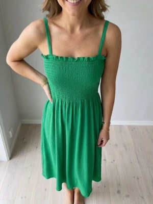 Grön sommarklänning med smock - Snygg grön klänning med smockad överdel och tunna axelband. Klänningen har en volangkant upptill och är tillverkad i ett lätt, luftigt material. Perfekt för varma dagar och enkel att styla med accessoarer.