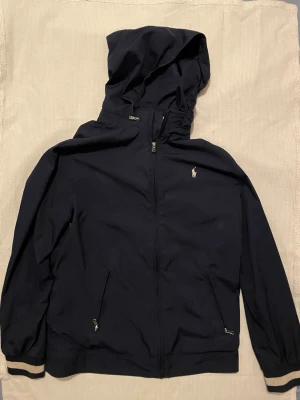 Marinblå vindjacka från Ralph Lauren - Snygg marinblå vindjacka från Ralph Lauren med huva och dragkedja framtill. 