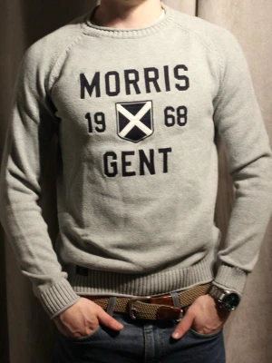 Grå tröja Morris Gent 1968 - Snygg grå tröja från Morris med trycket 'MORRIS GENT 1968' och ett skotskinspirerat emblem på bröstet. Tröjan har rund hals, ribbade muddar och är tillverkad i mjukt stickat material. Perfekt för dig som gillar klassisk men sportig stil.