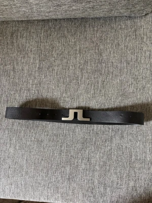 Svart skärp från J.Lindeberg i skinn - Stilrent svart skärp från J.Lindeberg i äkta skinn med en snygg silverfärgad JL-logga som spänne. Perfekt accessoar för att lyfta din outfit med en modern touch. Tillverkat i Italien och passar midjemått 30-31 tum.