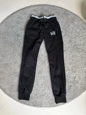 Svarta mjukisbyxor från Lounge - Svarta mjukisbyxor från Lounge med vit logga på vänster ben och elastisk midja med Lounge-tryck. Byxorna har dragsko i midjan, muddar vid benslut och är tillverkade i mjukt material för maximal komfort.