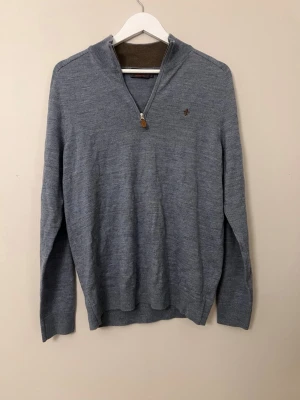 Grå merinoulltröja med zip från Morris - Säljer en stilren grå stickad tröja från Morris i 100% merinoull. Tröjan har halvzip med läderdetalj på dragkedjan, diskret logga på bröstet och ribbade muddar. Perfekt för lager-på-lager och snygg till jeans eller chinos.