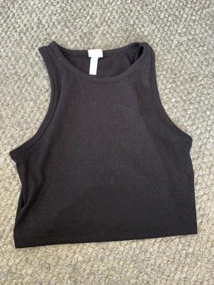 Svart ribbad croppad linnetopp - Säljer en svart croppad linnetopp från H&M med ribbad struktur. Toppen är ärmlös och har en hög halsringning som ger en sportig look. Perfekt att matcha med jeans eller kjol för en cool stil.