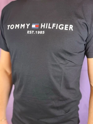 Svart t-shirt från Tommy Hilfiger - Säljer en svart t-shirt från Tommy Hilfiger med klassisk logotyp och text 'EST. 1985' på bröstet. T-shirten har korta ärmar och är tillverkad i mjuk bomull. Perfekt för dig som gillar stilrena och ikoniska plagg.