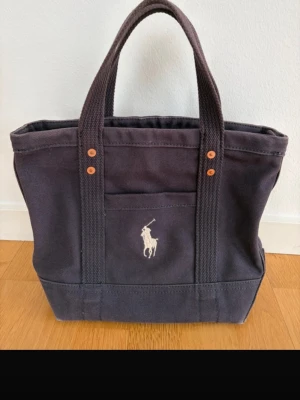 Ralph Lauren Tore bag mörkblå canvas  - Polo Ralph Lauren i kraftig canvas med klassisk broderad logga på framsidan. Väskan har två rejäla handtag. Perfekt för att bära allt du behöver under dagen.