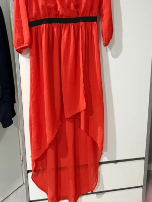 Röd långklänning med omlott - Säljer en röd långklänning med omlott-design och svarta detaljer i midjan. Klänningen har trekvartsärmar och är tillverkad i ett lätt och flödande material som ger en snygg siluett. Perfekt för dig som vill ha något färgstarkt och stilrent.