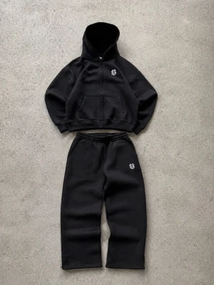 Svart loungewear-set med hoodie - Mysigt svart loungewear-set med hoodie och matchande byxor. Hoodien har dragkedja, fickor och en vit logotyp på bröstet. Byxorna har elastisk midja och samma vita logotyp på benet. Perfekt för chill dagar hemma. Materialet ser ut att vara mjukt och fleeceliknande.