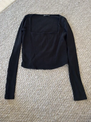 Svart ribbad topp från Zara - Trendig svart ribbad topp från Zara med lång ärm och fyrkantig urringning. Toppen är croppad och har en slim passform som framhäver siluetten. Perfekt att styla med både jeans och kjol.